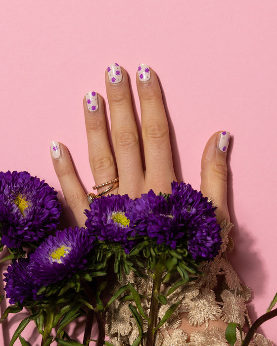Hand faced flat on a pink background with purpl flowers laying over it showing fingers with flower pattern nail art gel nail stickers